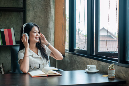 A young woman wearing headphones, sitting at a desk with a book and coffee, enjoying music in a...