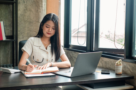 Focused woman writing notes at her desk, working on a laptop in a stylish home office setting.