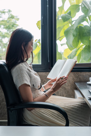 A woman sits in a comfortable chair reading a book by the window, with lush greenery visible outside.の写真素材