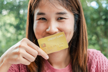 A woman smiles while holding a gold credit card, set against a lush outdoor background, symbolizing financial freedom and lifestyle.の写真素材