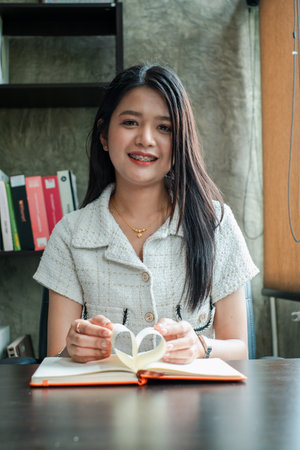 A young woman with long hair smiles while reading a book in a cozy office environment, featuring a bookshelf in the background.の写真素材