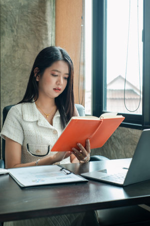 A young woman sits at a desk in a modern office, reading a book with a laptop nearby, illuminated by...