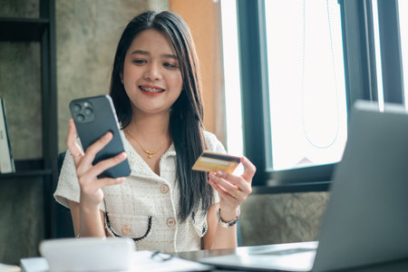 Smiling woman using smartphone and credit card for online shopping at home, sitting by laptop near window.の写真素材