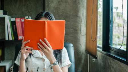 A woman sits in a modern office, reading a book by the window, surrounded by stylish decor and natural light.の写真素材