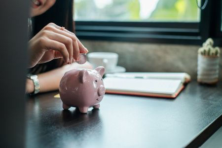 Close-up of a woman inserting a coin into a pink piggy bank on a desk, symbolizing savings and financial planning.の写真素材