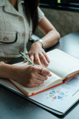 Professional woman taking notes in a notebook with financial charts on a desk, showcasing business and finance concepts.の写真素材