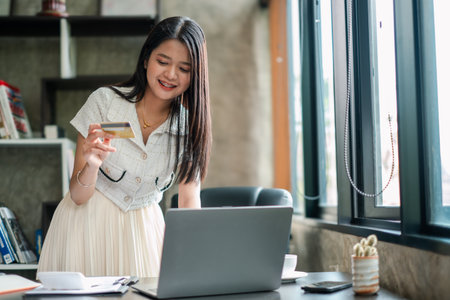 Smiling woman using laptop and credit card for online shopping in a modern home office setting.の写真素材