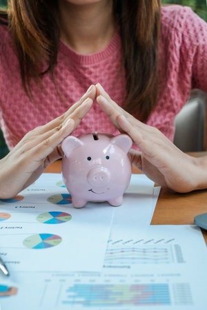 Close-up of a woman shielding a piggy bank with hands, surrounded by financial documents and colorful charts.の写真素材
