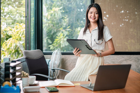 Smiling young woman in a modern office holding a tablet, with a laptop and documents on the desk, surrounded by greenery.の写真素材