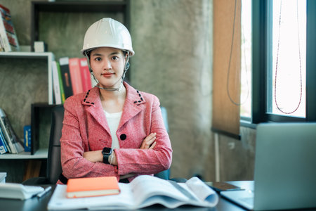 Female engineer in a pink blazer and hard hat, confidently seated at a desk in a modern office setting.の写真素材