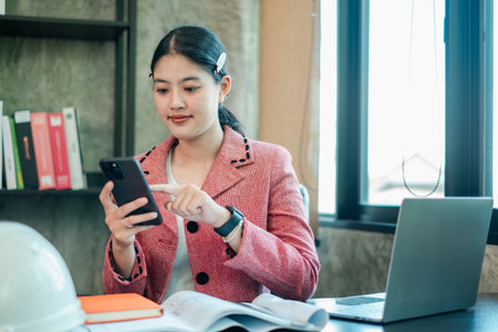 A young woman in a pink blazer uses her smartphone in a modern office, with a laptop and books on the desk.の写真素材