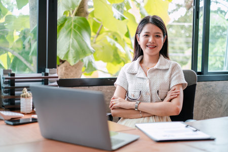Professional woman sitting at desk with laptop, smiling confidently in a bright office space with large windows and lush outdoor view.の写真素材
