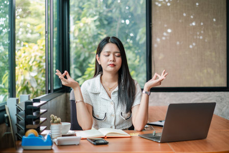 A young woman practices meditation at her desk in a modern office, surrounded by a laptop, notebook, and greenery outside the window.の写真素材