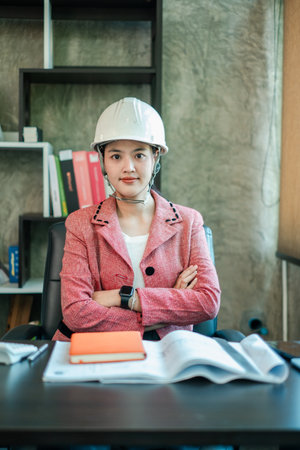 Professional woman engineer wearing a hard hat, sitting at a desk with documents, showcasing confidence and expertise in a modern office setting.の写真素材