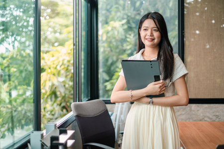 A young woman stands confidently in a modern office, holding a laptop. Large windows reveal lush greenery outside, creating a serene work environment.の写真素材