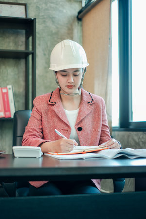 Female engineer wearing a hard hat, working diligently at a desk in a modern office setting.の写真素材