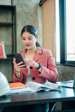 A young woman in a pink blazer uses her smartphone at a desk in a modern office environment.の写真素材