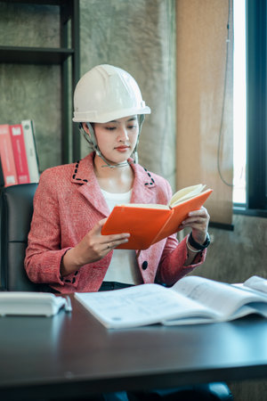 Woman engineer wearing a hard hat, reading a book at her desk in a contemporary office environment.の写真素材