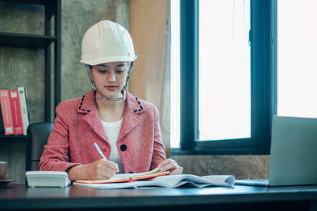 Professional female engineer wearing a hard hat, focused on work at her desk in a contemporary office setting.の写真素材