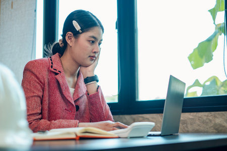 Young woman in a red blazer working on a laptop at a desk, with natural light streaming through a window in a modern office.の写真素材