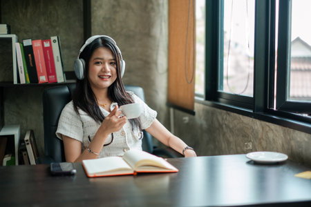 Smiling woman with headphones holding coffee cup, sitting at desk with open notebook in a bright office space.の写真素材