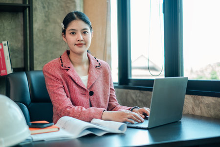 Businesswoman in pink blazer using laptop at office desk, surrounded by documents and a hard hat, near a large window.の写真素材