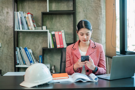 Businesswoman in a modern office using a smartphone, with a laptop and documents, showcasing a professional work environment.の写真素材