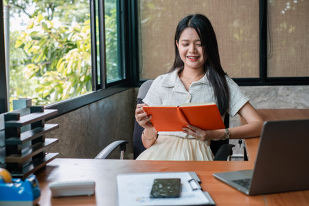 A young woman in a modern office setting, reading a book. The room is filled with natural light and...