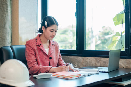 Businesswoman in a pink blazer working at a desk with a laptop and documents, showcasing a professional office environment.の写真素材