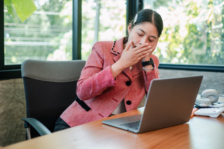 Tired businesswoman in a pink blazer yawning while working on a laptop in a bright, modern office environment.の写真素材