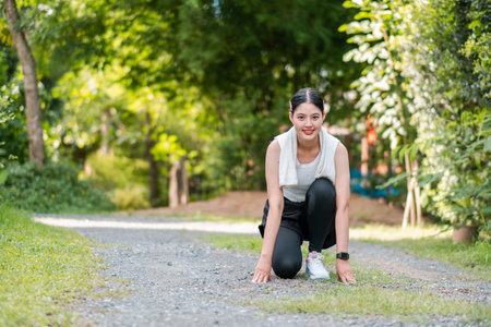 Woman in athletic wear kneeling on a gravel path, ready to run, with vibrant green trees and grass in the background.の写真素材