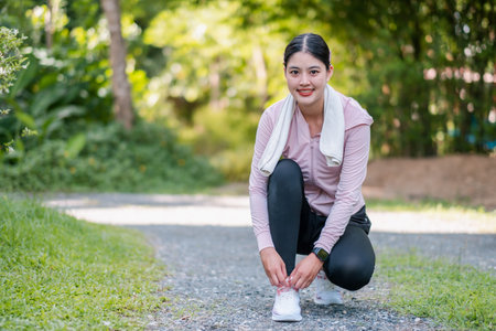 A woman prepares for a run by tying her shoelaces on a scenic park trail surrounded by greenery.