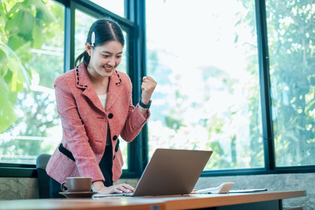 A joyful businesswoman in a pink blazer celebrates success while working on a laptop in a bright, modern office.の写真素材