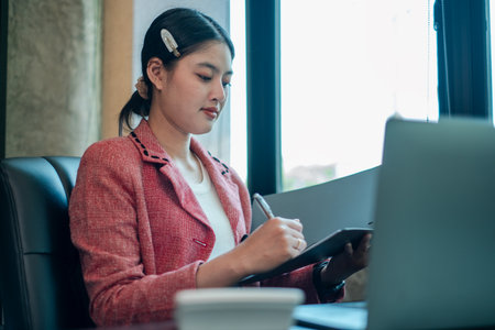 Focused woman in a red blazer writing notes at her desk, working on a laptop in a modern office setting.の写真素材