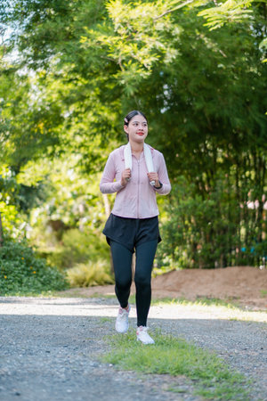 Woman jogging on a park path with a backpack, enjoying a sunny day amidst lush greenery.の写真素材
