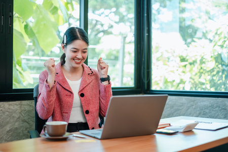 A joyful young woman in a pink blazer celebrates success while working on her laptop in a bright, modern office.の写真素材