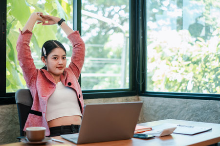 A young woman in a pink blazer stretches at her desk in a bright, modern office setting with a laptop and coffee.の写真素材