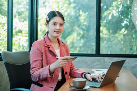 Businesswoman in a pink blazer working on a laptop, holding a credit card, in a bright office with...