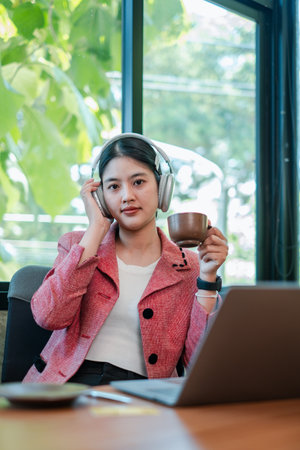Confident woman in a pink blazer using headphones and laptop, enjoying coffee in a bright, modern office environment.の写真素材