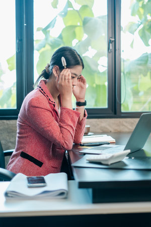 Professional woman in a pink blazer focused on work at her desk in a modern office environment.の写真素材