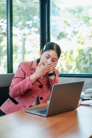 A tired businesswoman yawning at her desk with a laptop, highlighting workplace fatigue and the need for rest.の写真素材