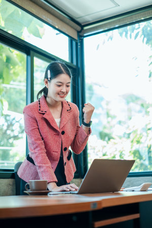 A young woman in a pink blazer celebrates success while working on her laptop in a bright, modern office space.の写真素材