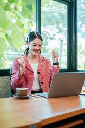 A joyful young woman in a pink blazer celebrates success while working on her laptop in a bright, modern office setting.の写真素材