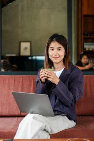 Smiling woman holding coffee cup, working on laptop in a cozy cafe. Relaxed and productive atmosphere.の写真素材