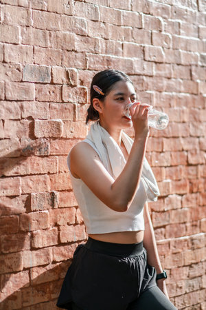 A young woman takes a break, drinking water after exercise, leaning against a textured brick wall, embodying health and fitness.の写真素材