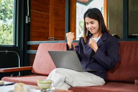A joyful young woman in a cafe, celebrating success while using her laptop, showcasing modern work and lifestyle.の写真素材