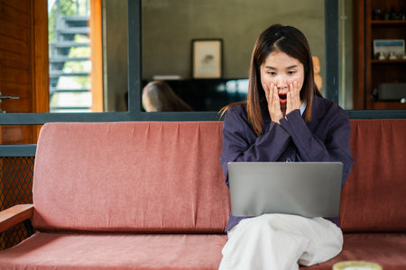 A young woman looks surprised while using a laptop on a sofa in a modern living room with wooden decor and large glass windows.の写真素材