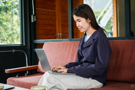 A young woman focused on her laptop in a stylish cafe with a warm, wooden interior and large windows.の写真素材