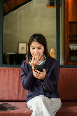 A young woman with headphones around her neck reacts to something on her smartphone while sitting on a sofa in a contemporary living room.の写真素材