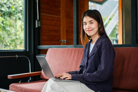 Smiling young woman using a laptop while sitting on a sofa in a stylish home office setting with natural light.の写真素材